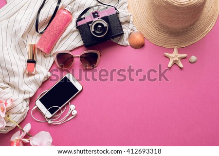 Overhead of essentials for modern student, teenager, woman. Top view of female fashion beach  accessories,straw hat, mobile phone with earphones, lipstick and body cream on bright pink background.