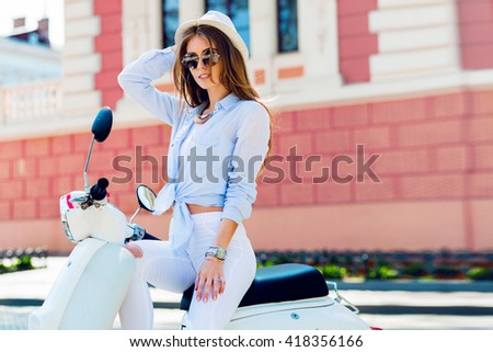 Close up lifestyle  image of young fashionable woman in casual outfit sitting on scooter   on the street.    Wearing blue shirt, white pants, trendy sunglasses. Tourist woman enjoying  holidays .