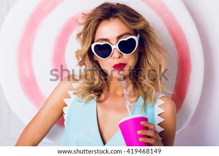 Sexy cute curly blonde  girl in sunglasses with beautiful skin and lips, posing in studio, drinking fruit juice or cocktail. Wearing vintage heart sunglasses, stylish blue leather top.