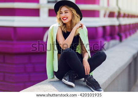 Outdoor lifestyle  close up portrait of happy blonde young woman in stylish  casual outfit sitting on bridge on the street. Pretty hipster girl having fun and enjoying  holidays.
