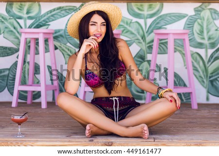 Summer fashion image of  young  woman in sexy beach bikini with tasty cocktail  sitting on wood floor in stylish  cafe .  Wearing boho accessories, straw hat, pink sunglasses.