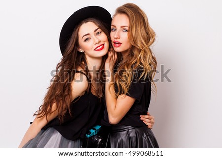 Close up fashionable  portrait  couple of two   cute girls best friends  smiling  and embracing  on white background .Perfect wavy hairstyle. Bright make up. White wall. Black stylish trendy hat.