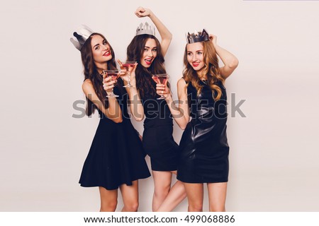Three beautiful young women celebrate hen-party and drinking cocktails on  white background. Best friends wearing black stylish evening dress, high heel shoes ,crown on head .