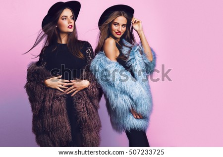 Positive fashion portrait of two girls, best friends posing indoor on bright pink background wearing winter stylish fluffy coat, black casual hat. Fashionable clothes.  Sisters walking.
