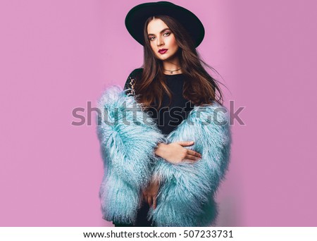 Close up indoor studio fashion portrait of gorgeous  woman in stylish winter fluffy  blue coat and black hat posing on bright pink background .Spa?e for text.