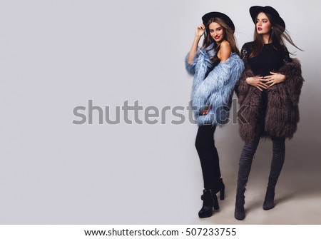 Positive fashion portrait of two girls, best friends posing indoor on grey background wearing winter fluffy coat, black casual hat. Fashionable clothes.  Sisters walking. Space for text. Full length .