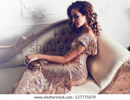 Beautiful stunning young lady in amazing glitter sequins dress  sitting on luxury armchair , holding  trendy evening hand bag. Bright red full lips, hollywood make up.