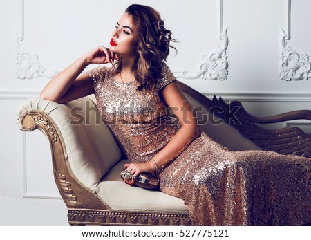 Beautiful stunning young lady in amazing glitter sequins dress  sitting on luxury armchair , holding  trendy evening hand bag. Bright red full lips, hollywood make up.