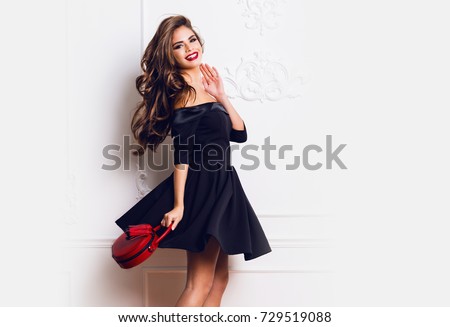 Amazing luxury seductive  woman in stylish  black  party dress posing on white  wall . Red hand bag . Bright smokey eyes make up, healthy wavy hairs. Fashion portrait. Celebrating   birthday  party.