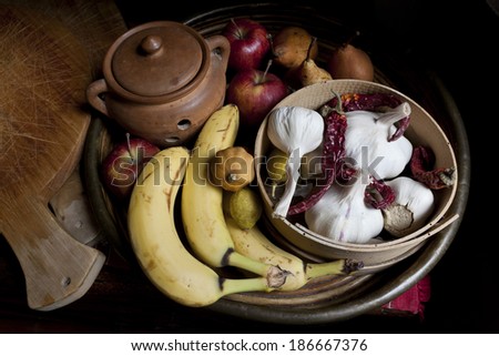 still life with whole fruits and vegetables like banana, lemon, apple, pear, red pepper, garlic on basket with wooden chopping board and ceramic pot in rustic background