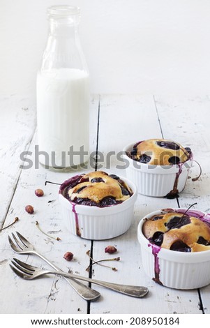 three french clafoutis with blueberries and cherries on ceramic ramekins on rustic white vintage background with milk glass bottle