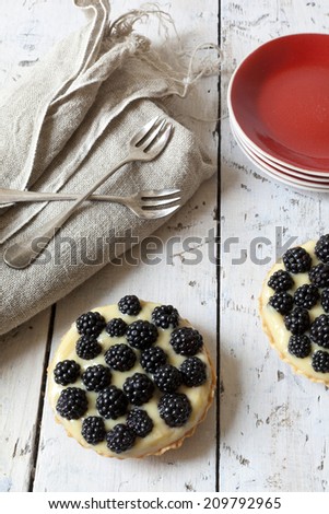 two blackberries tart with pastry cream on wooden table with cloth, fork and little plate