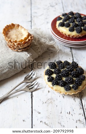 two homemade blackberries tart with pastry cream on wooden table with cloth, fork and little plate with tart waffles