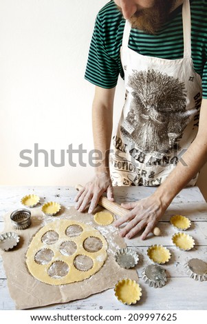 bearded young man with apron making shortcrust pastry for little tart on molds on rustic background with rolling pin and flour on wooden white table
