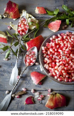 pomegranate grains on bowl with two silver spoon full of grains on table with pomegranate peel and leaves on rustic wooden table