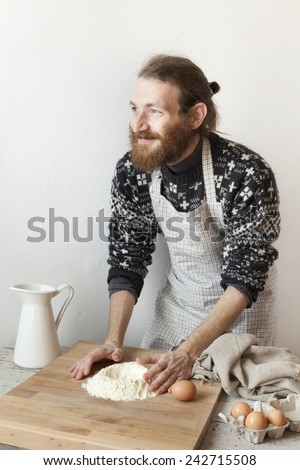 young bearded stylish man in the kitchen with apron making dough for pasta with white flour and eggs on wooden rustic board