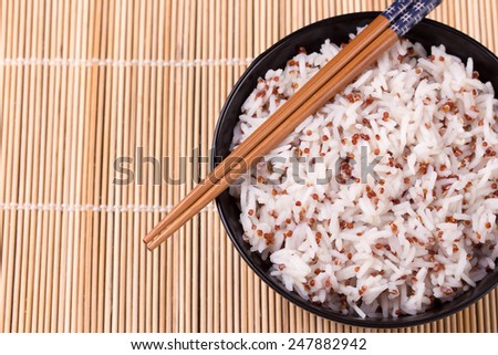 Black bowl of white steamed rice mixed with quinoa and chopsticks on bamboo mat. Copy space.