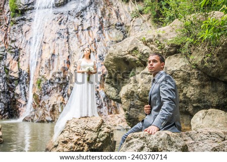 Portrait of the groom on the rock, and the bride on background near waterfall
