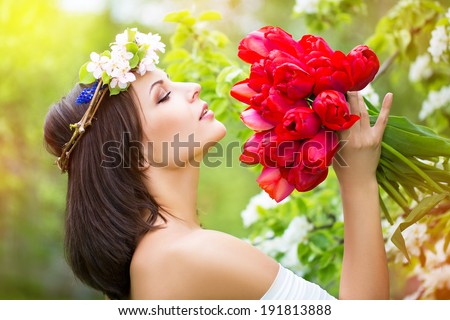 Portrait of a beautiful young woman in a wreath of spring flowers with a bouquet of red tulips in his hands outdoor in the flowered garden