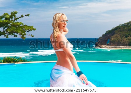 Young beautiful blonde girl in a white swimsuit with a waving plume of white cloth is next to the pool. Tropical sea in the background. Summer vacation.