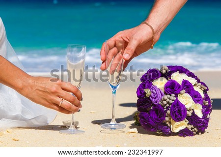 Wedding bouquet and two glasses of champagne on the sand. Male and female hands with wedding rings are drawn to the glass. Blue sea in the background.