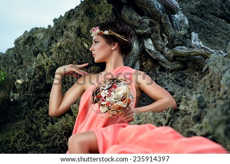 Young beautiful girl with a wreath in the sea theme dressed in pink fabric sitting on a rock and holding a bouquet of shells.