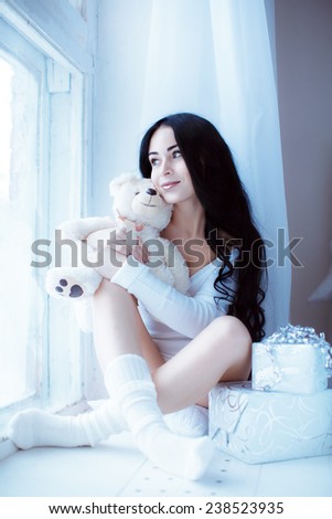 Young beautiful girl in a white sweater sits at the winter window with a toy bear in my hands and gift boxes. Christmas and new year concept.