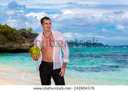 Happy handsome young male wearing in white shirt is standing on the tropical beach is holding coconuts under the sun on sunny summer day during holidays vacation.