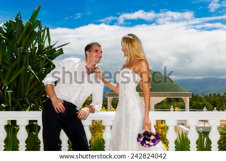 Happy bride and groom standing next to the stone gazebo amid beautiful tropical landscape. Sea, sky, flowering plants and palm trees in the background. Wedding and honeymoon concept.