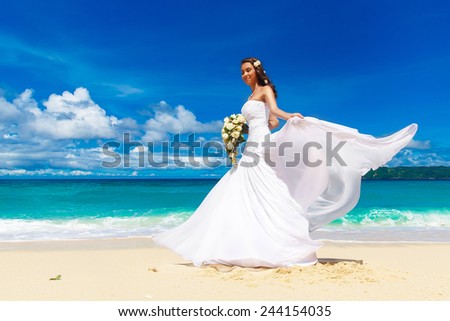 Beautiful brunette bride in white wedding dress with big long white train and with wedding bouquet stand on shore sea. Tropical sea in the background. Summer vacation concept.