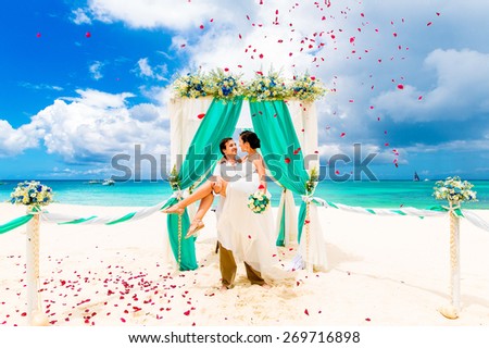 Wedding ceremony on a tropical beach in blue. Happy groom and bride under the arch decorated with flowers on the sandy beach. Rose petals fall from above. Wedding and honeymoon concept.