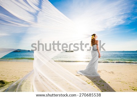 beautiful fiancee in white wedding dress and big long white train, stand on shore sea at sunset