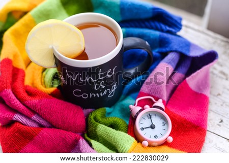 A mug with chalk writing Winter is coming with tea and lemon slice on it, wrapped in a multicolor rainbow scarf with tiny pink alarm clock set to 9 am