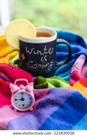 A mug with chalk writing Winter is coming with tea and lemon slice on it, wrapped in a multicolor rainbow scarf with tiny pink alarm clock set to 9 am