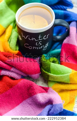 A mug with chalk writing Winter is coming with tea and lemon slice on it, wrapped in a multicolor rainbow scarf