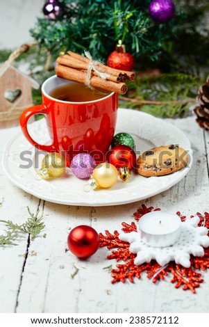 Red cup with tea for Christmas and New Year decoration with chocolate chips cookie , cinnamon sticks, colorful baubles with Christmas tree in background