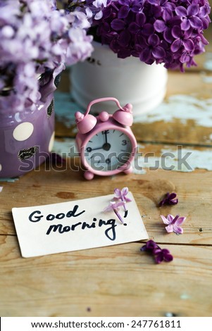 Two tone Lilac flowers in a ceramic pots white and purple, with pink vintage tiny alarm clock and a Good Morning note on a shabby wooden surface