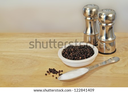 Two silver salt and pepper shakers with black peppercorns in a white bowl, sea salt and some spilled onto a wooden table.