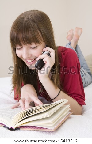 A teenager girl lying on a bed reading a book and talking on a mobile  phone at the same time