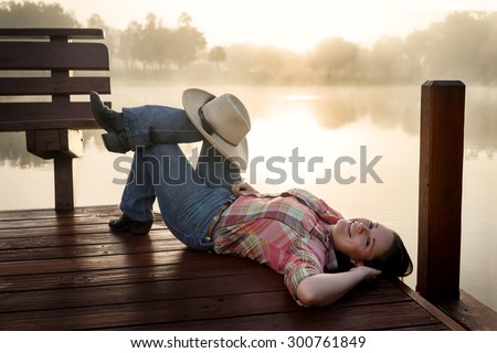 Girl woman lady with long dark brown hair lying down on a lake pond dock with reflections at sunrise or sunset in cowboy hat flannel shirt looking relaxed happy serene beautiful young  peaceful