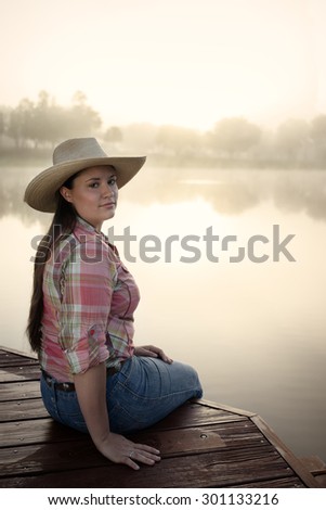 Cowgirl lady woman female wearing cowboy hat and flannel shirt with jeans leaning on country rural fence by a horse pasture paddock looking confident happy serene smart alone waiting watching patient