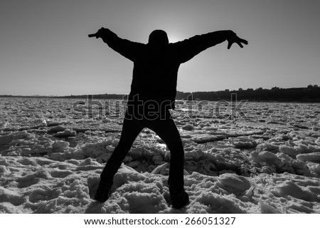 Black and white picture of the silhouette of a man in front of the sun holding the hands up with icy river background