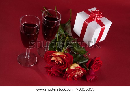 bouquet of red roses, white gift and two glasses of wine on a red background