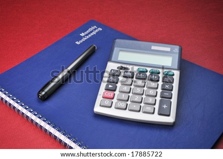 Business Books, Calculator, Expenses, Bookkeeping, Pen on top of  blue spiral bound monthly accounting income and expense reports with deep red background.