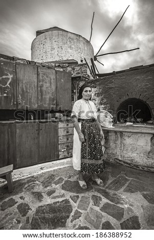 KARPATHOS, GREECE - SEPTEMBER 30, 2010: Woman in traditional clothes, stands in front of a stone traditional oven, with a windmill in the background, cooking for a tavern she runs with Greek food.