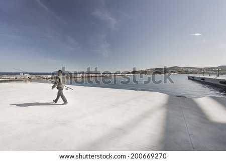 SYROS ISLAND, GREECE - MARCH 4, 2012:  A man walks towards a jetty, near the sea, holding a fishing pole, in order to spend some time alone, fishing. A  common hobby for those who live on an island.