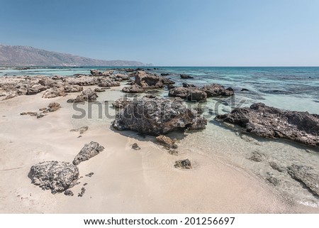 A sandy beach with some rocks inside a crystal clear, turquoise calm water, and a clear blue sky, during summer time, in the daylight, located in the island of Crete, Greece, near Elafonisi beach.