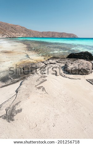 A sandy beach with some rocks inside a crystal clear, turquoise calm water, and a clear blue sky, during summer time, in the daylight, located in the island of Crete, Greece, near Elafonisi beach.