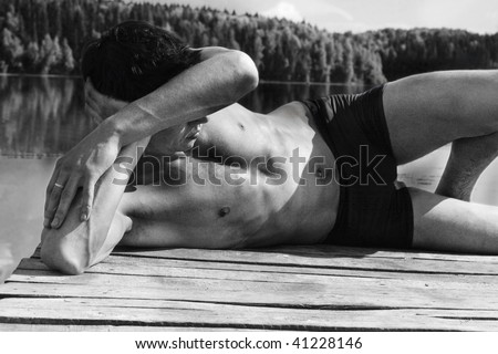 Young man in swimming trunks is lying against lake / black and white picture