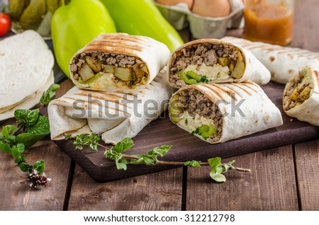 Breakfast burrito on wood board and wood table, eggs, pepper, potatoes and meat inside